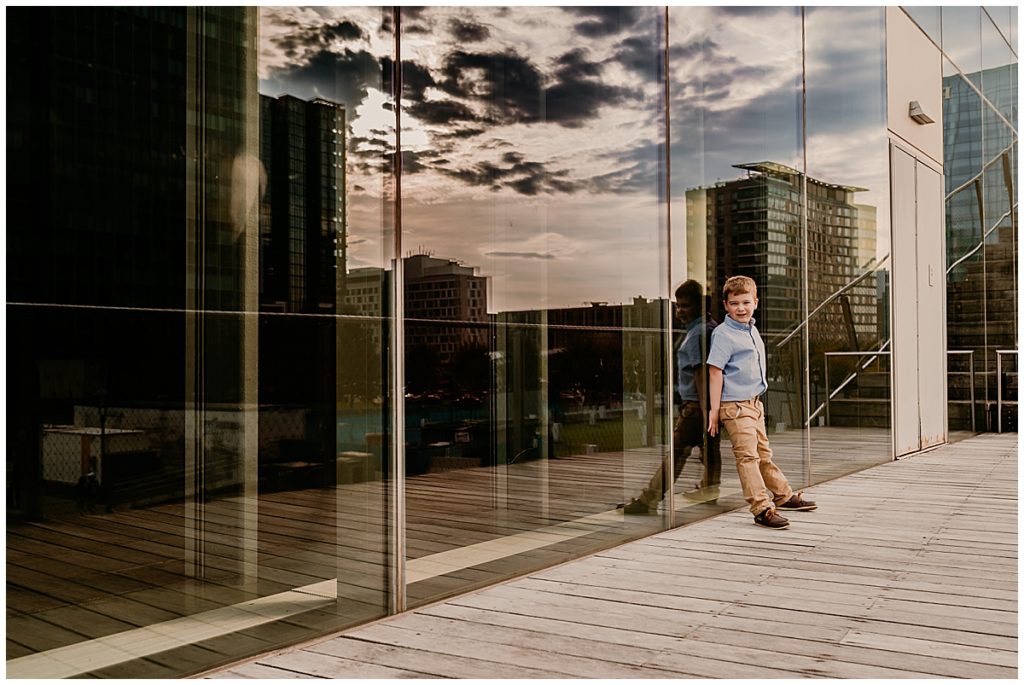 Boy standing against the glass wall of a building, with the boston buildings reflecting. 
