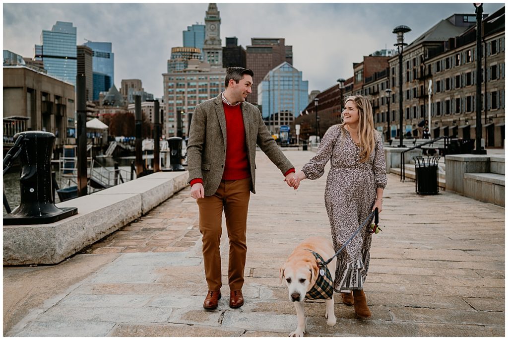 Man, woman and dog walking along paved path with Boston in background.
