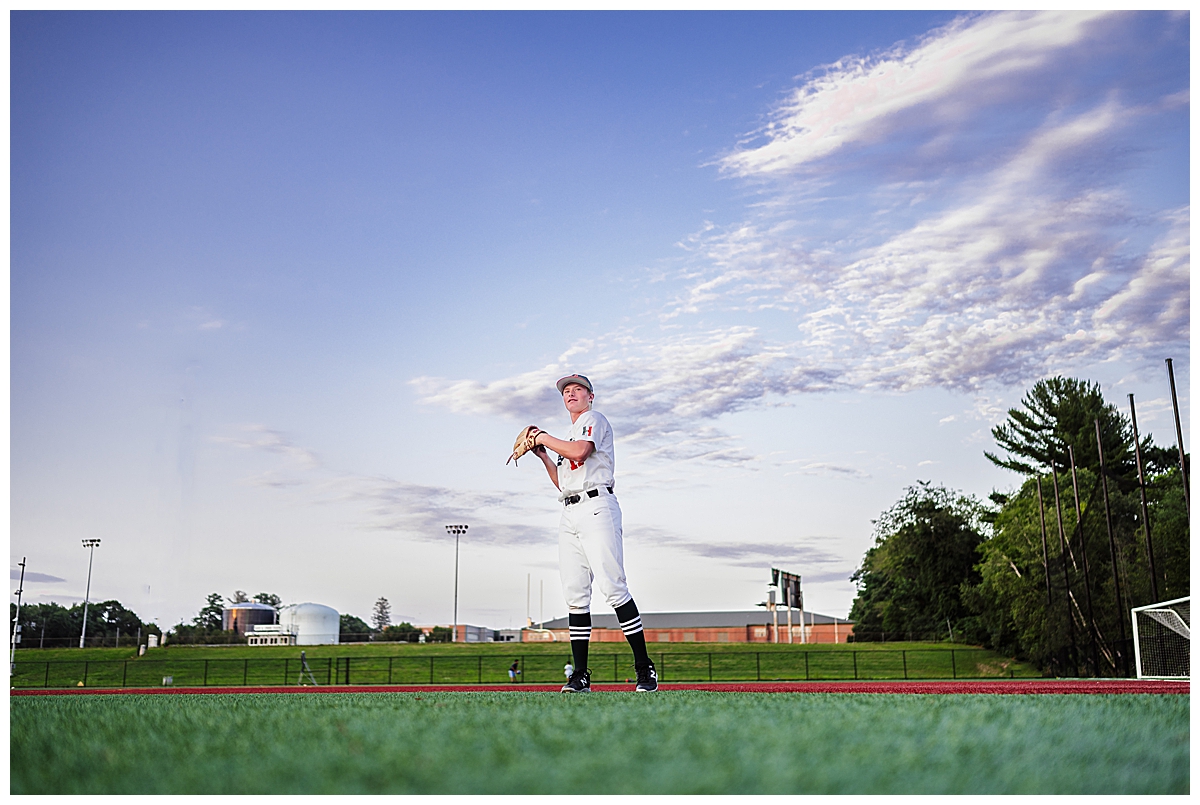 Boy standing on a baseball diamond holding a baseball with clouds and a blue sky behind him