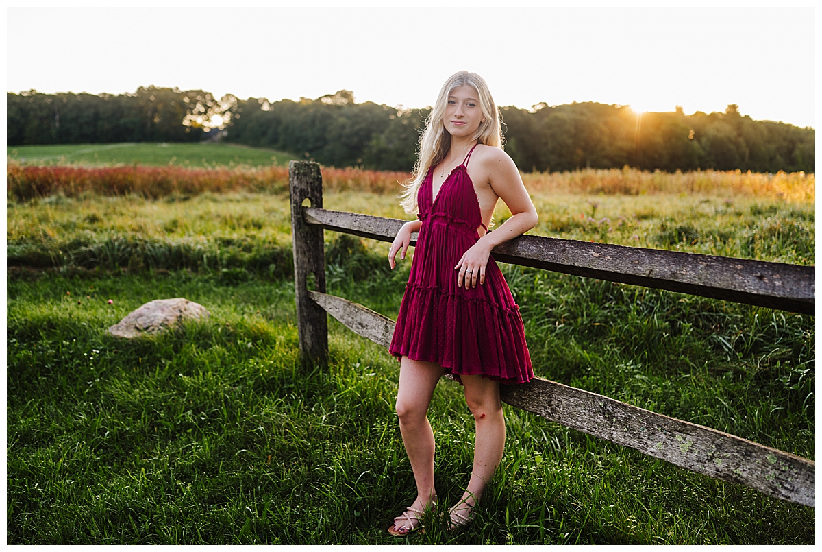 Girl wearing a red dress standing in a field against a fence posing for senior photographer.