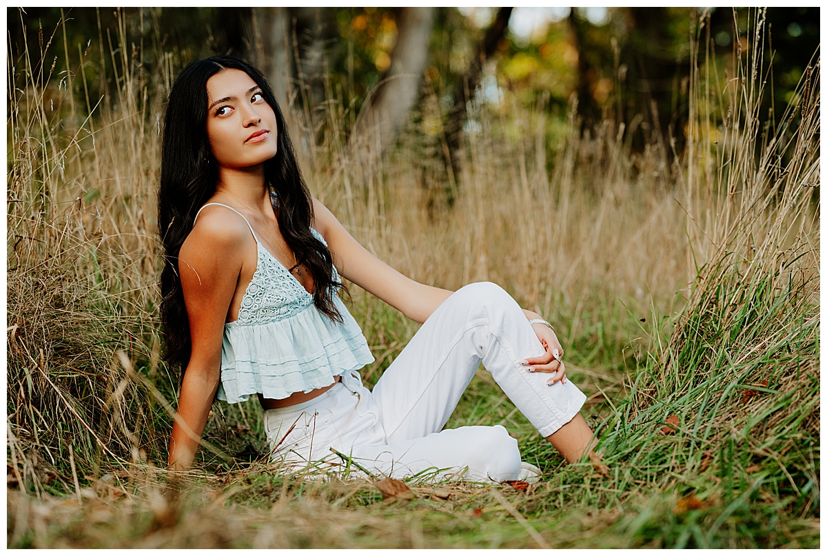 Girl wearing a blue tank top and white jeans sitting in a field looking over her back shoulder.