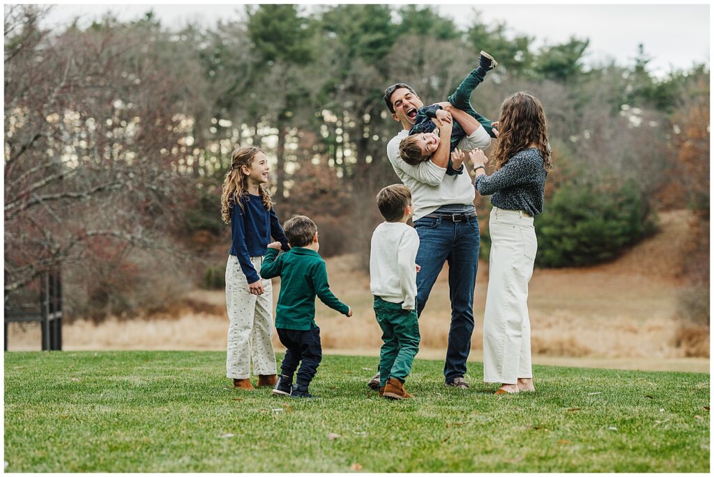 A family standing in a field wearing khakis with the father holding the boy in the air and the rest of the family smiling and laughing