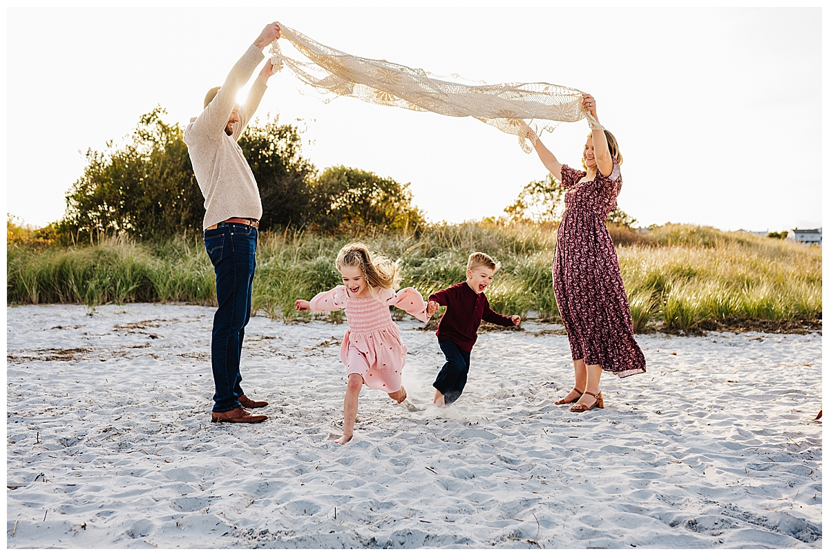 A joyful family of four walks barefoot on a sandy beach during sunset. The mother, wearing a red dress, holds her son's hand while the father, dressed in a brown sweater, carries their young daughter in a yellow dress. They are all smiling as they stroll together with the sky painted in soft blue and golden hues. A scenic coastal home and boats in the harbor are visible in the background.