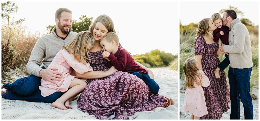 A family of four sits closely together on a sandy patch surrounded by tall grass. The mother, wearing a purple floral dress, hugs her two children—a daughter in a pink dress and a son in a burgundy sweater