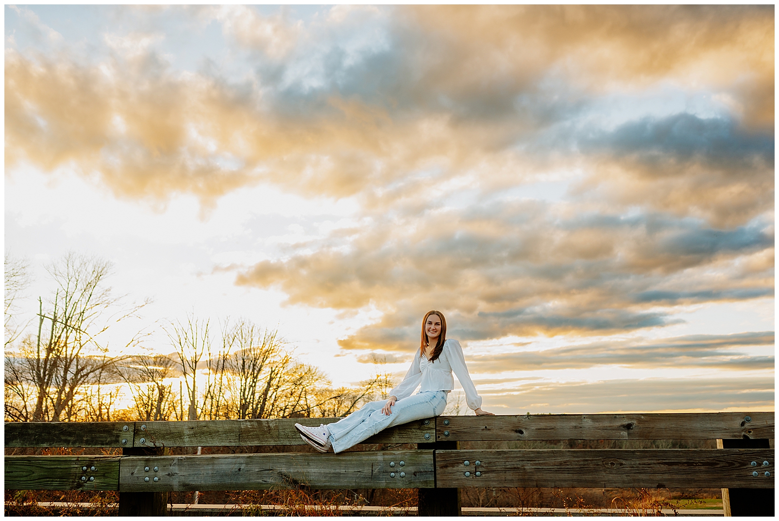 Girl wearing white shirt sitting on a fence with a bright sky and clouds behind her