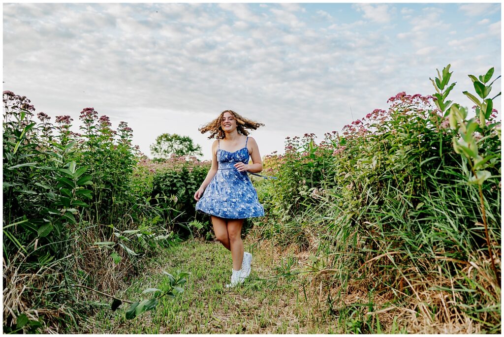 Senior girl with curly hair wearing a blue dress and sneakers twirling in a wild flower field during her senior photo session. 