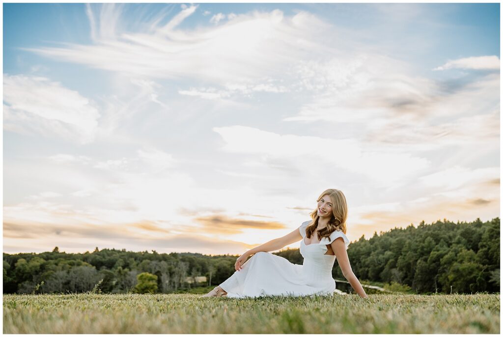 High school senior sitting in a grassy field with a sunset sky behind her during a relaxed outdoor senior photo session.
