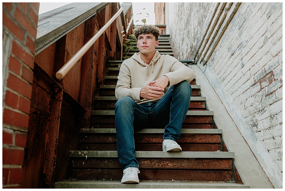 High school senior boy sitting on metal stairs between brick and painted walls in downtown Milford during an urban senior photography session.
