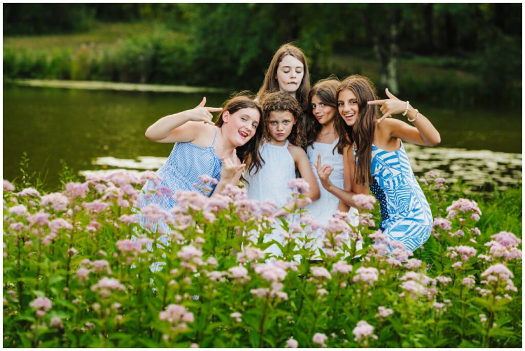 Five girl cousins making silly faces and posing together in a pink wildflower field next to a pond, candid extended family photography
