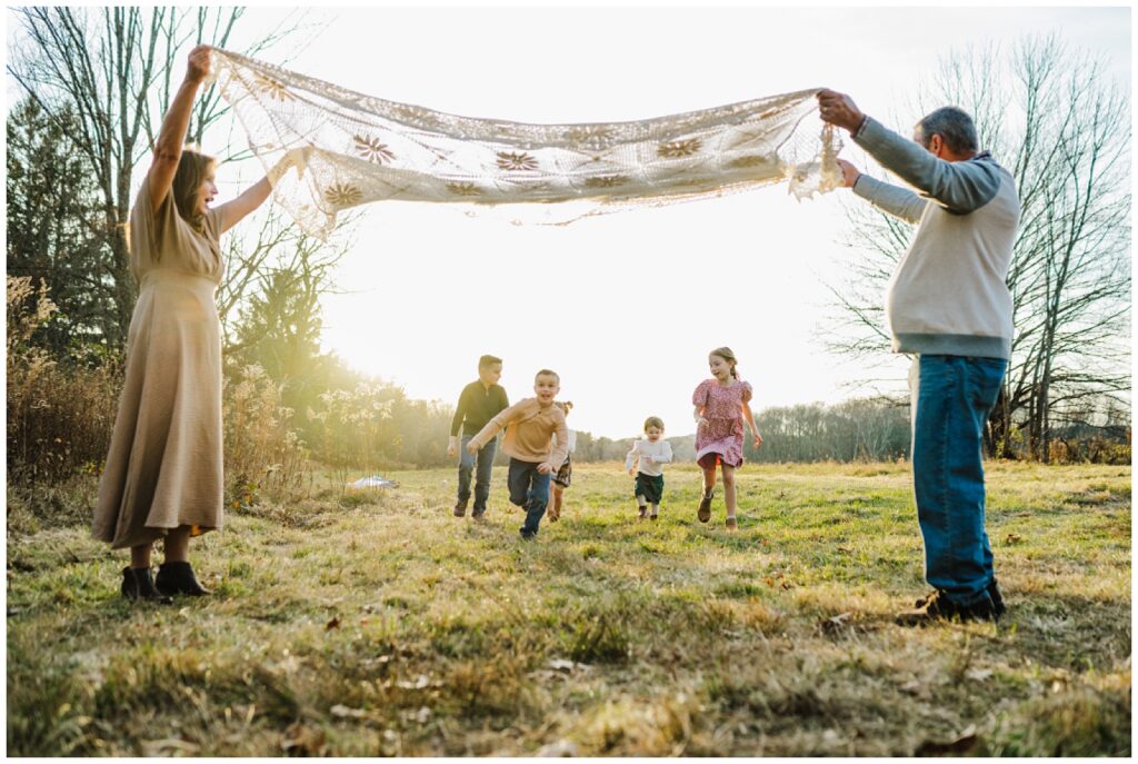 Grandparents holding a lace blanket overhead while grandchildren run through at golden hour in an open New England field in Hopkinton Massachusetts