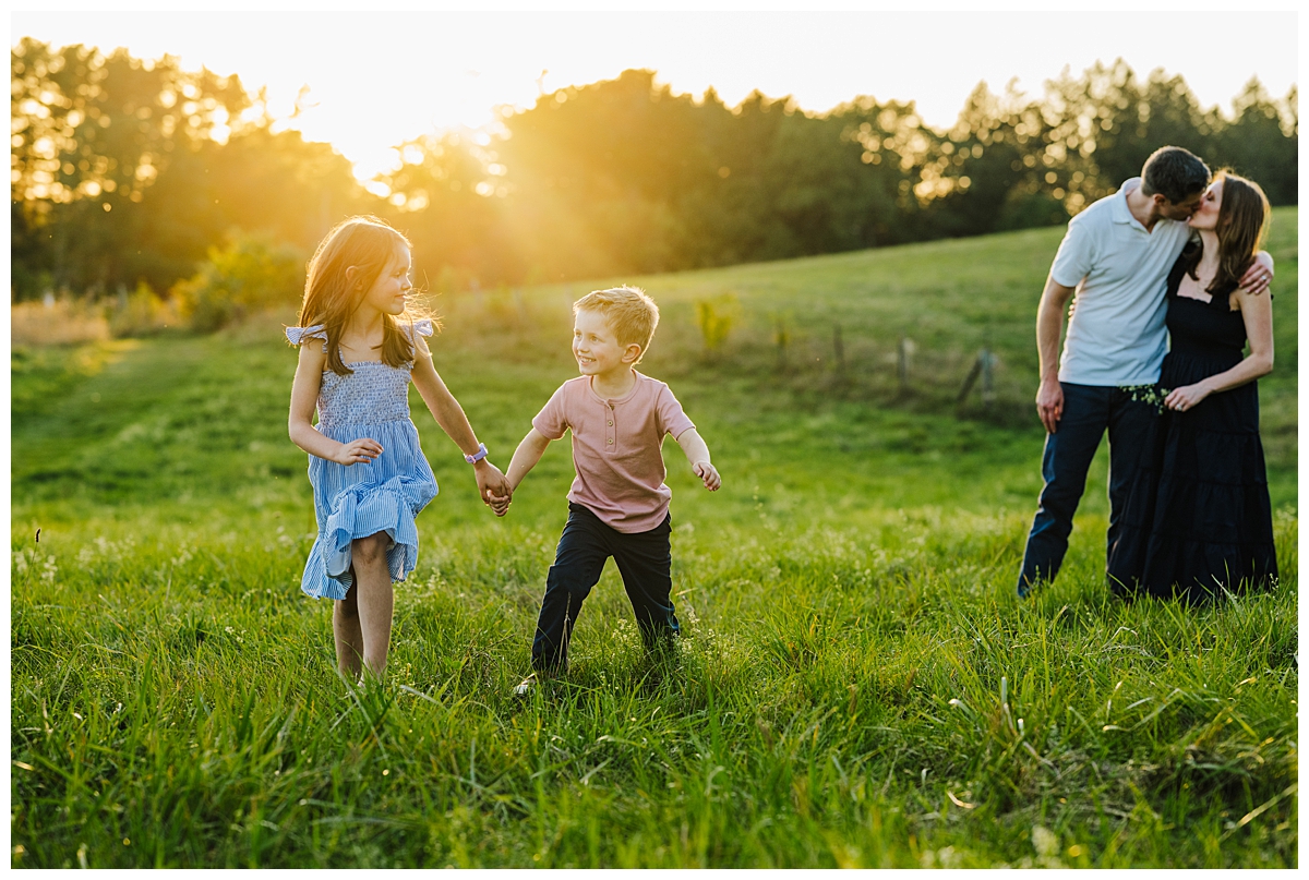 Sister and brother holding hands running through a sunlit field while parents kiss in the background in MetroWest Boston.
